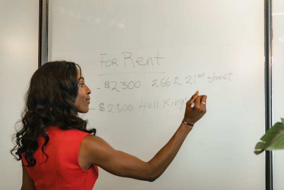 A woman with long, dark, wavy hair, wearing a sleeveless red top, is writing on a whiteboard with a black marker. The whiteboard displays handwritten notes related to property rental costs, including amounts of $2300 and $2500, as well as addresses on 21st Street and Kings. The woman is inside a room with neutral lighting, and part of a potted plant is visible in the foreground on the right. This scene appears to be set in an office or meeting space, potentially during an initial consultation or planning session for a house removal or moving service, as provided by Crystal Palace Man and Van. The focus is on her action of writing, capturing the process of discussing or planning a home relocation, with the whiteboard contents indicating cost estimates and addresses related to moving logistics, consistent with the service category of removals.