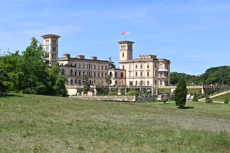 A large, ornate historic building with a cream-colored stone facade, featuring multiple towers, decorative battlements, and a central flagpole flying a Union Jack flag. The structure is set within well-maintained gardens with neatly trimmed shrubs, small trees, and a terraced lawn area that slopes gently downward. The building appears to be a castle or stately mansion situated in a spacious, open park with a clear blue sky overhead. The setting suggests a scenic location, possibly in the UK. Addressing home relocation and furniture transport, this exterior image captures the grandeur of a residence that could be part of a house moving project by Crystal Palace Man and Van, highlighting the importance of careful packing and logistics in moving large properties.