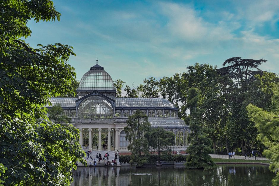 A large glass Victorian-style conservatory with a domed roof and elaborate metal framework, situated in a park setting with lush green trees surrounding it. The conservatory is visible behind a body of water, reflecting parts of the structure. The foreground includes leafy branches and trees, with some people walking on pathways near the building. The scene is illuminated by natural daylight under a partly cloudy sky, and the environment suggests a peaceful, outdoor setting associated with gardens and leisure areas. As part of a house or property relocation, Crystal Palace Man and Van may be involved in transporting or handling valuable or fragile items like the conservatory, highlighting their expertise in furniture transport and moving logistics.