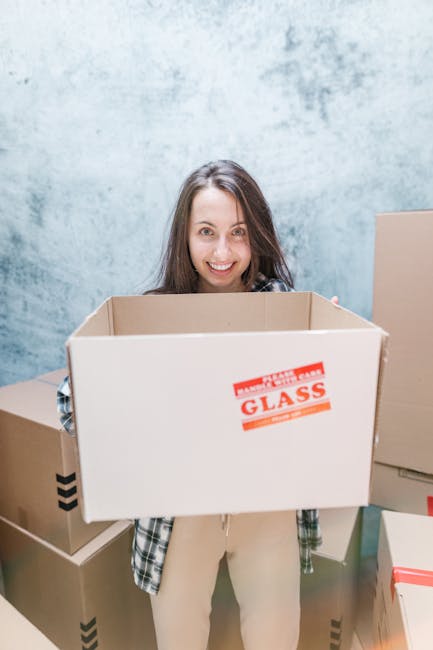 A woman with long brown hair smiling at the camera while holding an empty cardboard moving box labeled 'Glass' in a room with a blue mottled wall. Surrounding her are additional cardboard boxes of various sizes, some sealed with packing tape and others open, placed on the floor and adjacent surfaces. The scene appears to be part of a home relocation or packing and moving process, possibly prepared for transportation by Crystal Palace Man and Van, a professional removals service in SE19. The boxes are arranged in a way that suggests the stage of packing or loading, with the woman engaging in the process of furniture transport or house removals, highlighting the logistical steps involved in a same-day move to or from Crystal Palace.