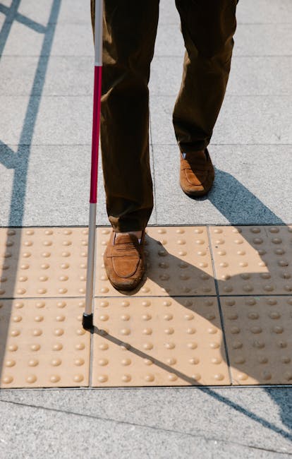 Close-up of a person wearing brown leather shoes and dark trousers, standing on tactile paving tiles outside a building entrance. The individual is holding a pink walking cane with a black tip, indica